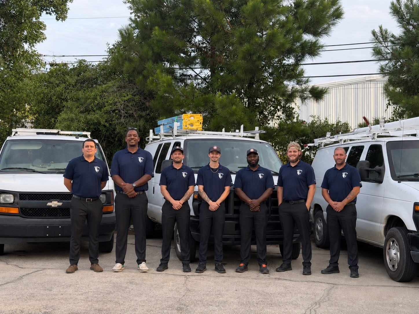 Seven uniformed Fogle & Sons Electric technicians standing together in front of the company&rsquo;s white service vans